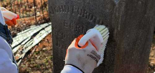 A worker using a brush to clean engraved letters on a dark granite headstone during restoration