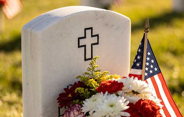 A white marble headstone cross decorated with flowers and a US flag in a cemetery